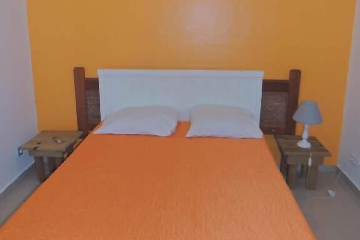 A room with an orange bedspread, a white headboard, two white pillows, a nightstand with a lamp, and an air conditioning unit on the wall.