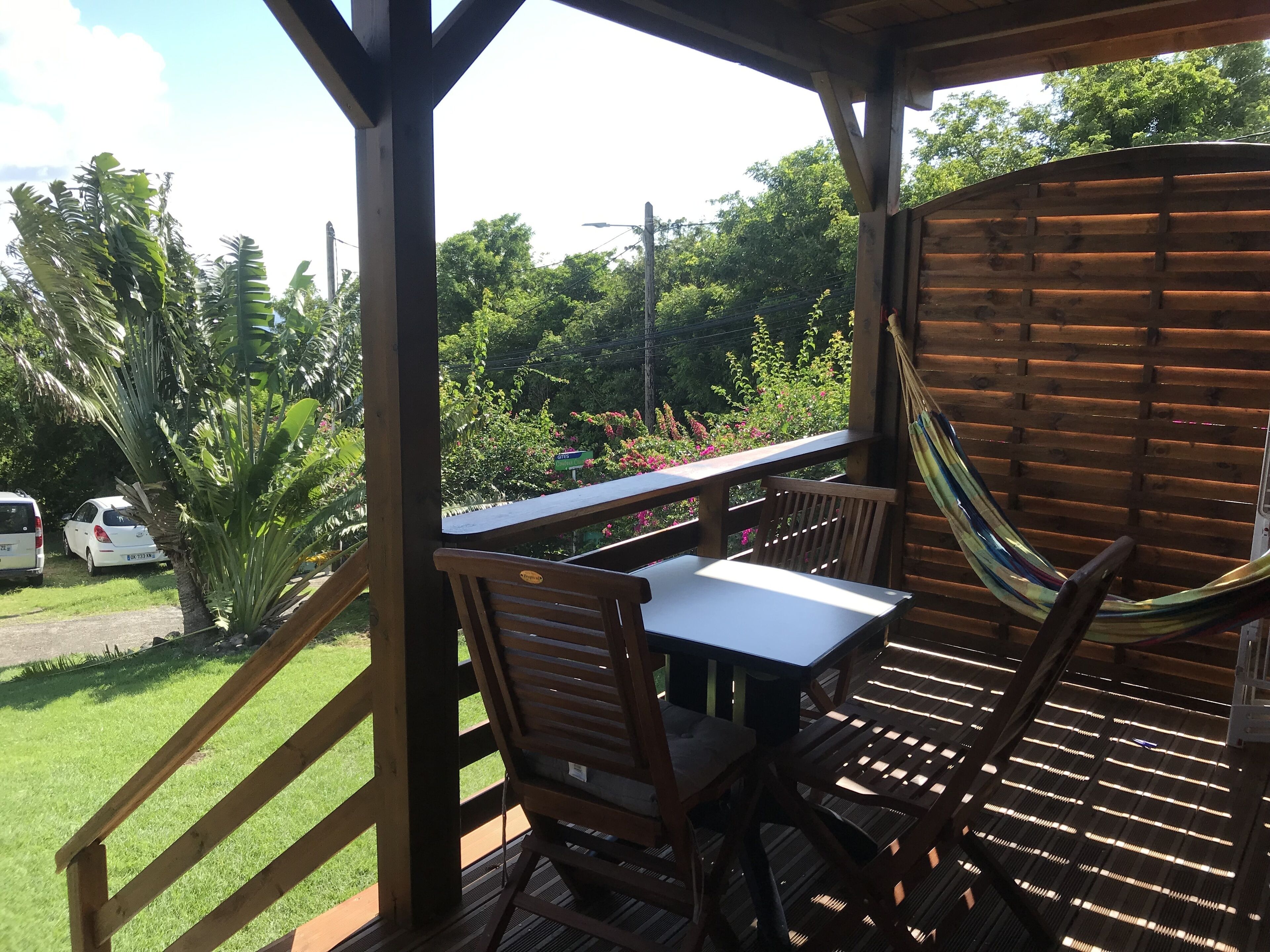 Una terraza de madera con una mesa y sillas, una hamaca y vistas a un jardín exuberante y coches aparcados.
