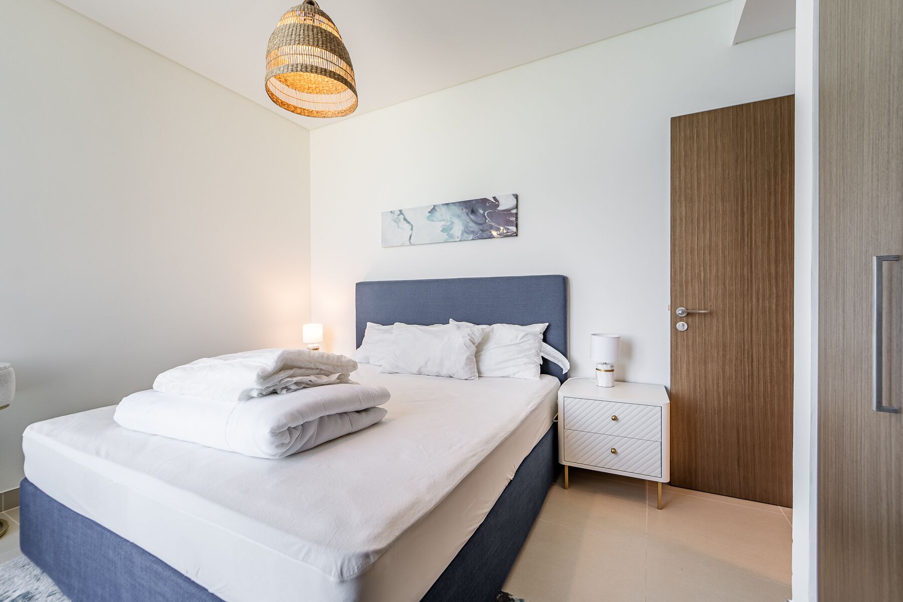 A neatly made bed with white linens, a bedside table with a lamp, and a wooden door.
