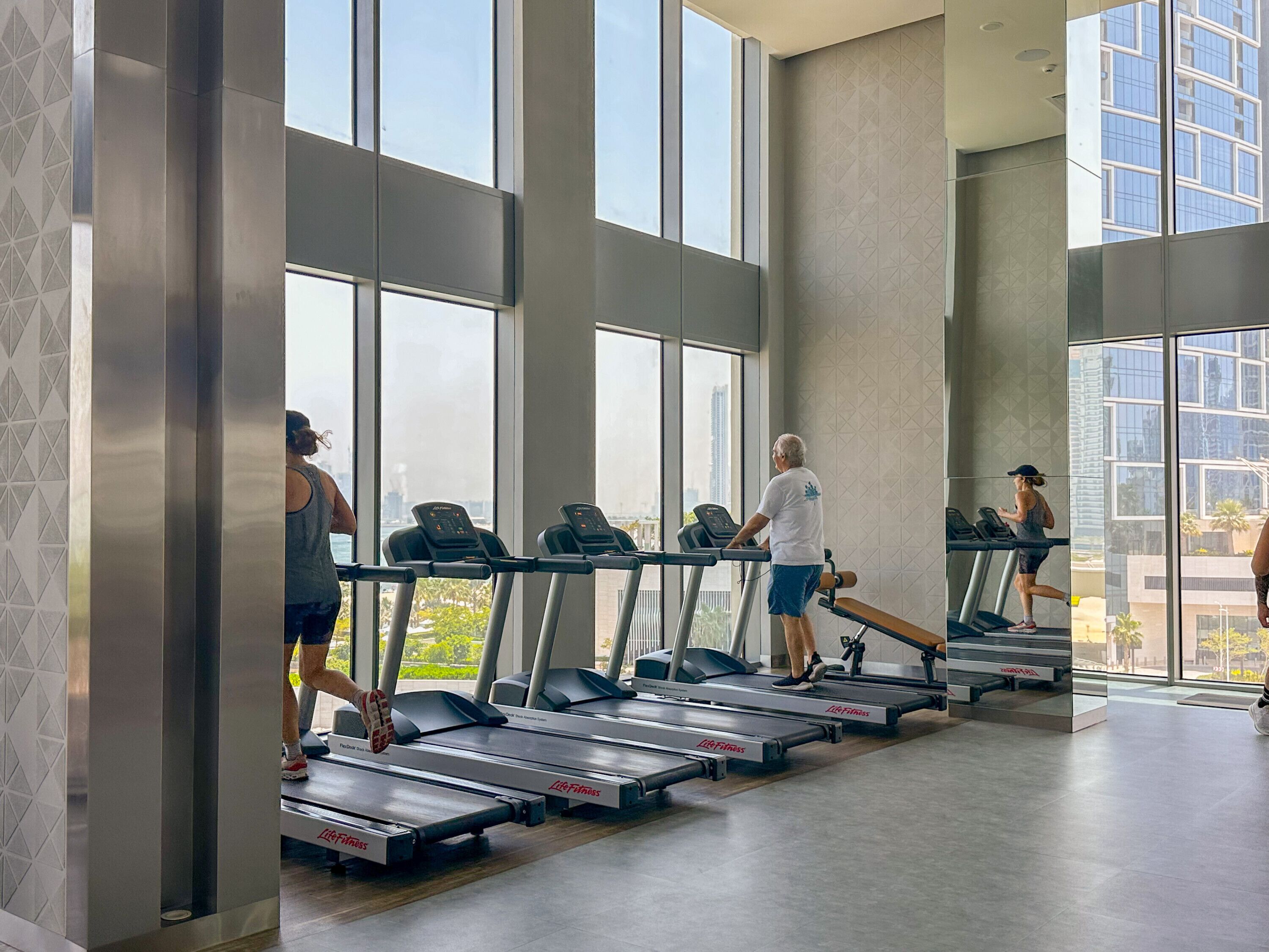 A modern gym with treadmills aligned in rows, large windows, and a view of the cityscape.