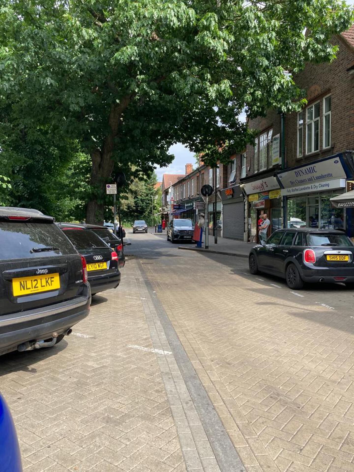 A street lined with parked cars and a row of shops, including a dry cleaning service.