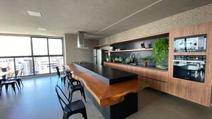 A modern kitchen with a central island, built-in appliances, and a large window offering a city view. - Apartamento na Ponta Verde - Maceio Alto Padrao Super Aconchegante (Ponta Verde Maceió)