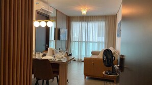 A hotel room with a dining area, a sofa, a standing fan, and a large window with curtains. - Apartamento na Ponta Verde - Maceio Alto Padrao Super Aconchegante (Ponta Verde Maceió)