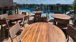 A rooftop pool area with round wooden tables and wicker chairs, surrounded by glass railings and a clear view of the city skyline. - Apartamento na Ponta Verde - Maceio Alto Padrao Super Aconchegante (Ponta Verde Maceió)