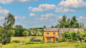 A yellow house with a sloped roof, surrounded by lush greenery and a clear blue sky. - Beili Glas Cottage (Llanfihangel Ystern Llewern)