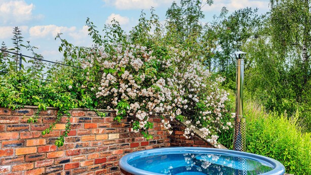A wooden hot tub next to a brick wall with flowering plants. - Beili Glas Cottage (Llanfihangel Ystern Llewern)