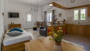 A modern, well-lit room with a bed, bedside table, and a dining table with a vase of flowers. There is a kitchen area with wooden cabinets and a window. - Cozy and comfortable Cycladic home to enjoy hospitality in Dryos of Paros Island (Paros)