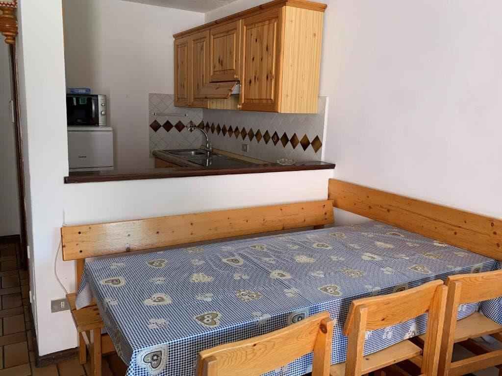 Una pequeña cocina con armarios de madera, un microondas y un lavamanos. Hay un banco de madera con mantel a cuadros.