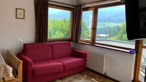 Quarto com sofá vermelho, cadeira de madeira, mesa com suporte para prato, radiador e televisão.