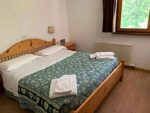 A single bed with a wooden headboard, a bedside table, a wall-mounted lamp, and a window with curtains. - Holiday apartment with mountain view (Cavalese)