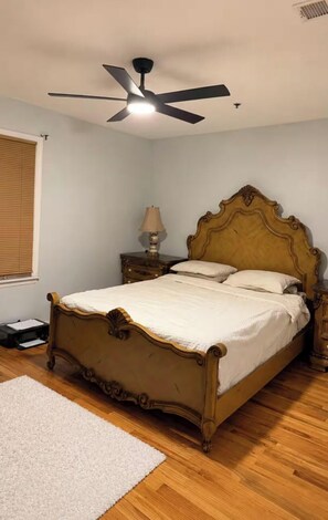 A bedroom with a wooden bed, a ceiling fan, a bedside table with a lamp, and a window with blinds. - Spacious LuxuryEscape 3 Bedrooms 2 Baths •           15 Mins to NYC.FREE PARKING (Jersey City)