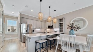 A modern kitchen with a central island, stainless steel appliances, pendant lighting, and a dining area with white chairs and a vase of flowers.