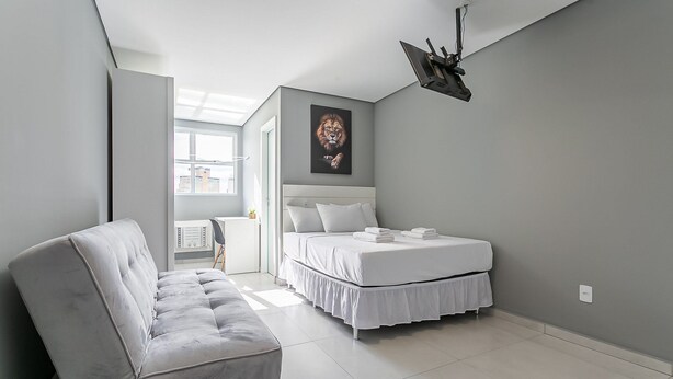 A modern bedroom with a grey couch, a bed with white bedding, and a ceiling-mounted projector. - New studio near Beiramar Shopping #HKS25 (Florianópolis)