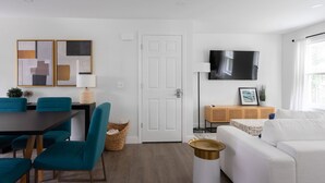 A modern living room with a dining area, a white door, a mounted TV, and a large window. - 3 BR in the Heart of Middletown, Minutes from Beach! (Middletown)