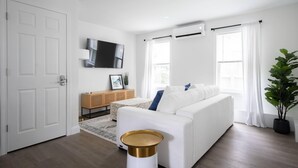 A modern living room with a white sofa, a small side table, a mounted TV, and a potted plant. - 3 BR in the Heart of Middletown, Minutes from Beach! (Middletown)