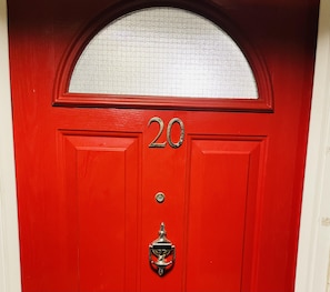 Puerta roja con el número 20, un panel de vidrio en la parte superior y un aldaba metálica en la parte inferior.