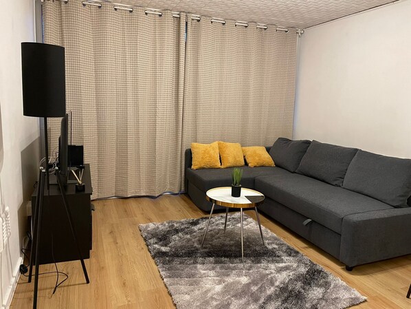 Una sala de estar con un sofá gris, una mesita redonda con una planta y un soporte negro para altavoz.