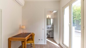 A compact room with a wooden table and chair, a small lamp, a white sink, and a large window offering a view of greenery.