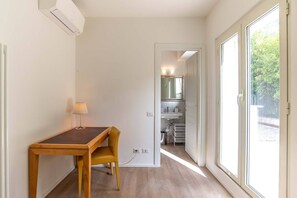 Quarto compacto com uma mesa e cadeira de madeira, uma luminária pequena, uma pia branca e uma janela grande com vista para vegetação.