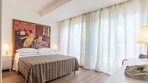 A hotel room with a bed, two bedside lamps, a painting on the wall, and a window with sheer curtains.