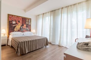 Quarto de hotel com cama, duas luminárias de cabeceira, um quadro na parede e uma janela com cortinas leves.
