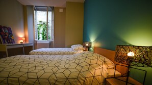 Une chambre avec deux lits, une fenêtre donnant sur un espace verdoyant, un radiateur et une lampe sur une table de chevet.