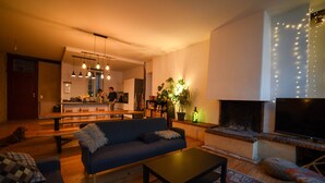 Un salon chaleureux avec un canapé, une table basse et une cheminée. On aperçoit une cuisine au fond, où une personne est en train de cuisiner.