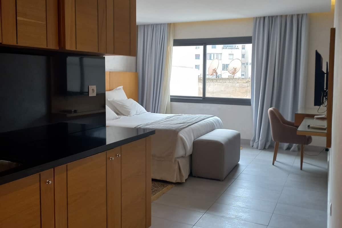 Une chambre d’hôtel moderne dotée d’une kitchenette, d’un lit avec une grande fenêtre, d’un coin salon et d’une télévision.