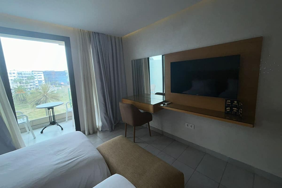 Une chambre d’hôtel équipée d’un lit, d’un bureau, d’une chaise, d’une télévision et d’un balcon offrant une vue sur les immeubles et les palmiers.