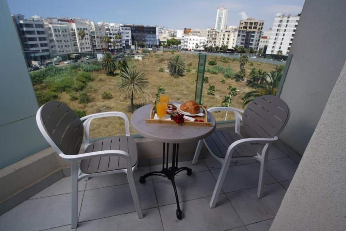 Un balcon avec deux chaises et une table, donnant sur un paysage urbain composé d’immeubles et d’une zone verdoyante.