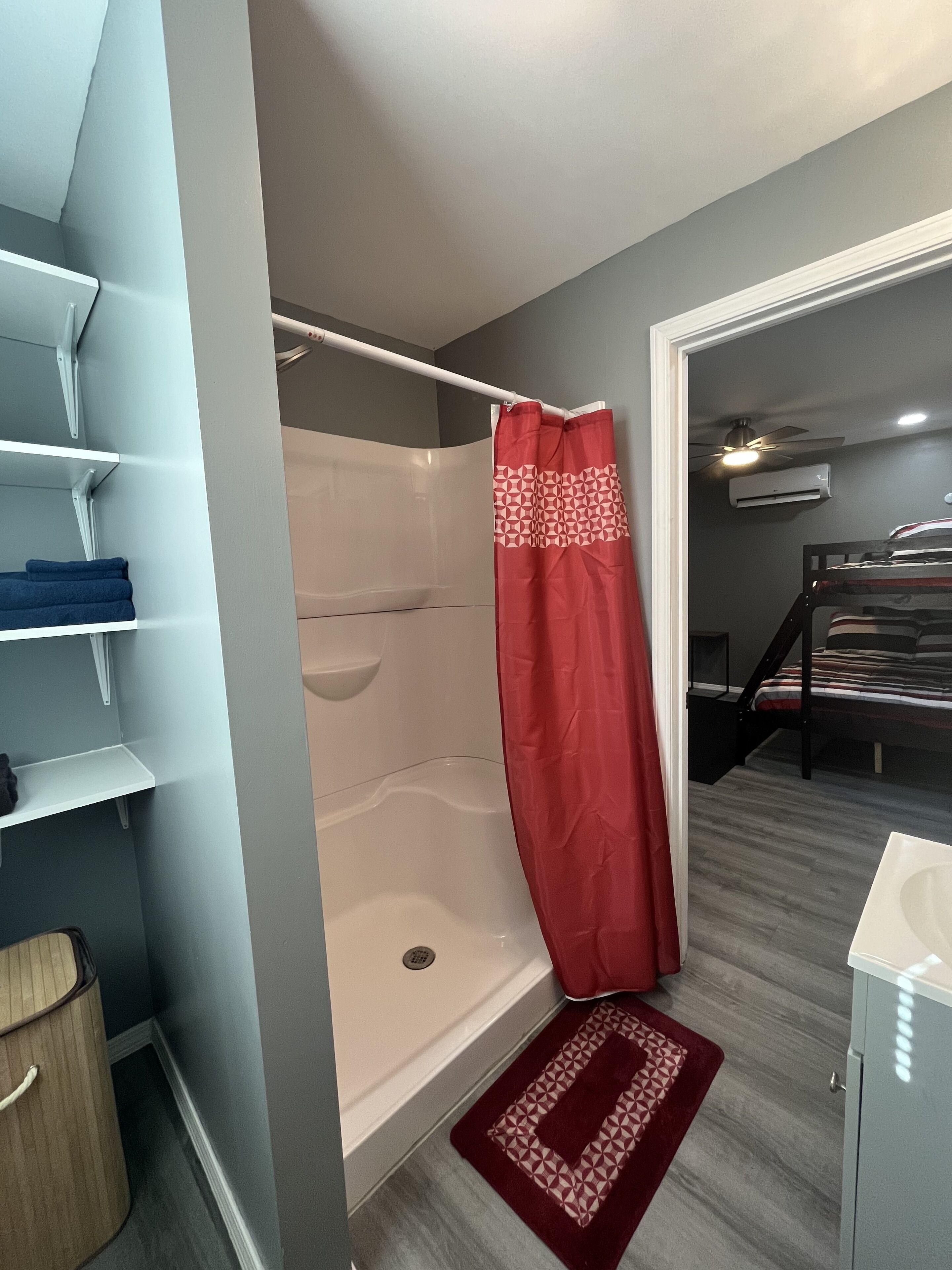 Een badkamer met een douche, een rood douchegordijn, een houten emmer, een witte wastafel en een rood tapijt.