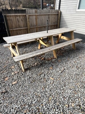 A wooden picnic table with benches, set on gravel ground, surrounded by a wooden fence and a building. - Luxury ATV lodging with beautiful view, sleeps 11. (Welch)