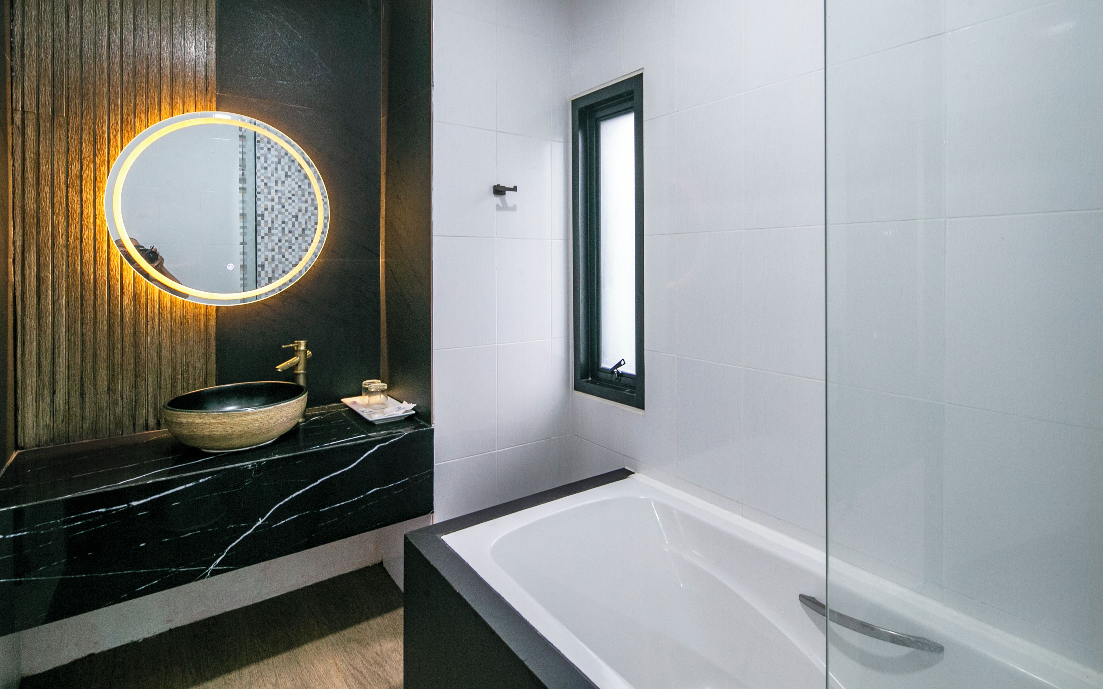 Une salle de bain avec un meuble-lavabo en marbre noir, un miroir rond avec un éclairage jaune, un mur recouvert de panneaux en bois, une baignoire blanche et une fenêtre.