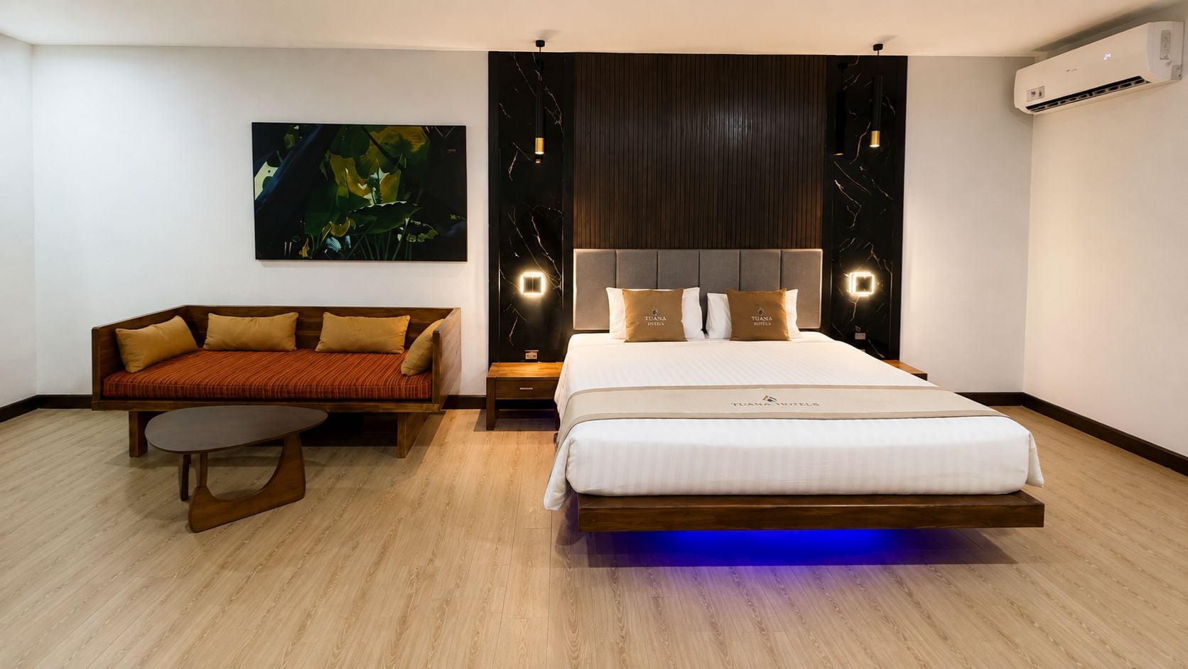 A modern hotel room with a bed, a sofa, a nightstand, and a wall-mounted air conditioner.