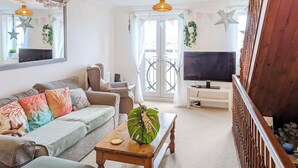 Ein Wohnzimmer mit Sofa, Couchtisch und Fernseher. Ein Balkon mit Geländer und Blick ins Freie ist vorhanden.