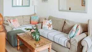Ein Wohnzimmer mit einem beige farbenen Sofa, einem Holztisch und einem grünen Hocker. Auf dem Sofa liegen Zierkissen und ein Blumenvasen.