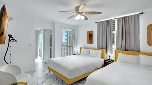Habitación de hotel con dos camas, ventilador de techo, ventana grande con cortinas, televisor montado en la pared y una pequeña mesa con una planta.