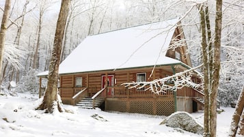 Uma cabana de madeira com telhado coberto de neve, rodeada por árvores numa floresta nevada.