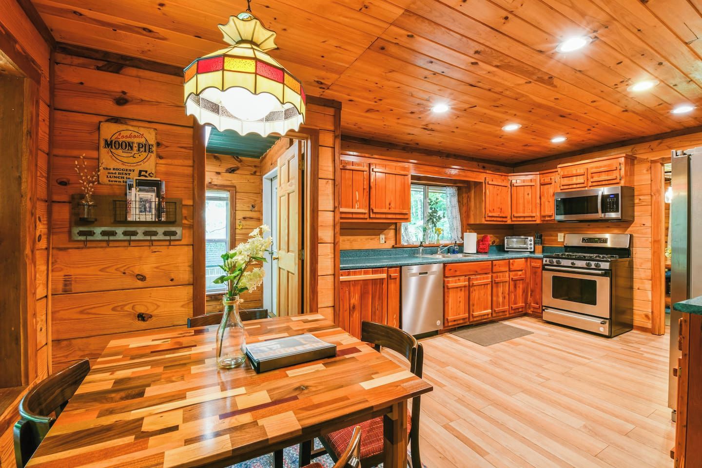 A wooden cabin interior with a dining table, a chandelier, wooden cabinets, a refrigerator, and a stove.