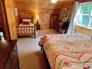 A wooden cabin bedroom with a bed, a dresser, a nightstand, and a window. - Creekside log cabin near Lake Chatuge & Blue Ridge (Hayesville)