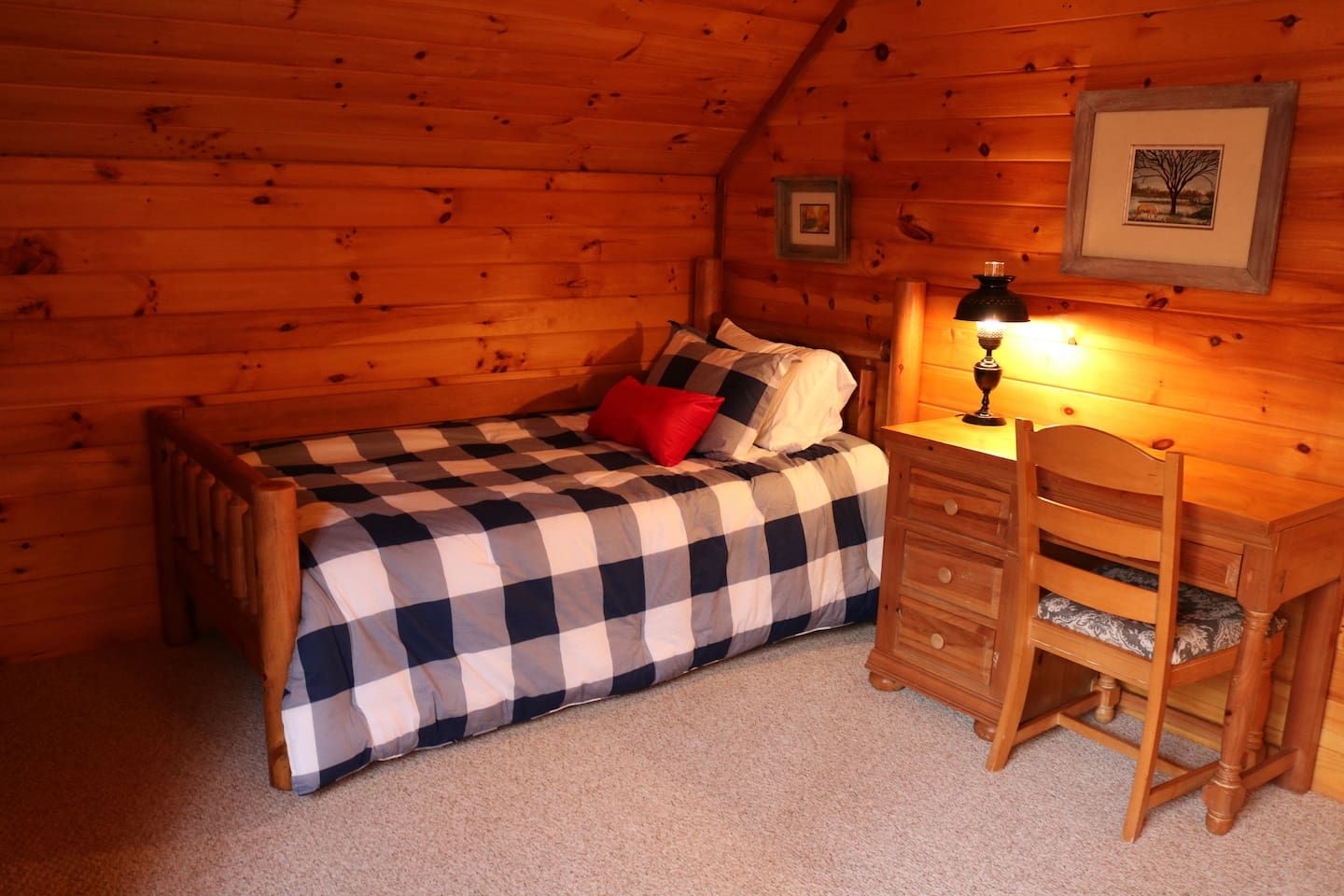 Un dormitorio de cabaña acogedor con colcha de cuadros, un cojín rojo, un escritorio de madera con cajones y una lámpara.
