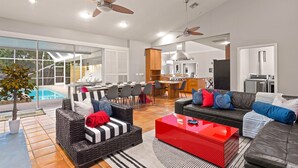 A spacious living area with a dining table, sofas, and a red coffee table. There is a kitchen in the background and a pool outside. - Next to IMG Golf! Heated Pool, Spa, Game Room! (Bradenton)