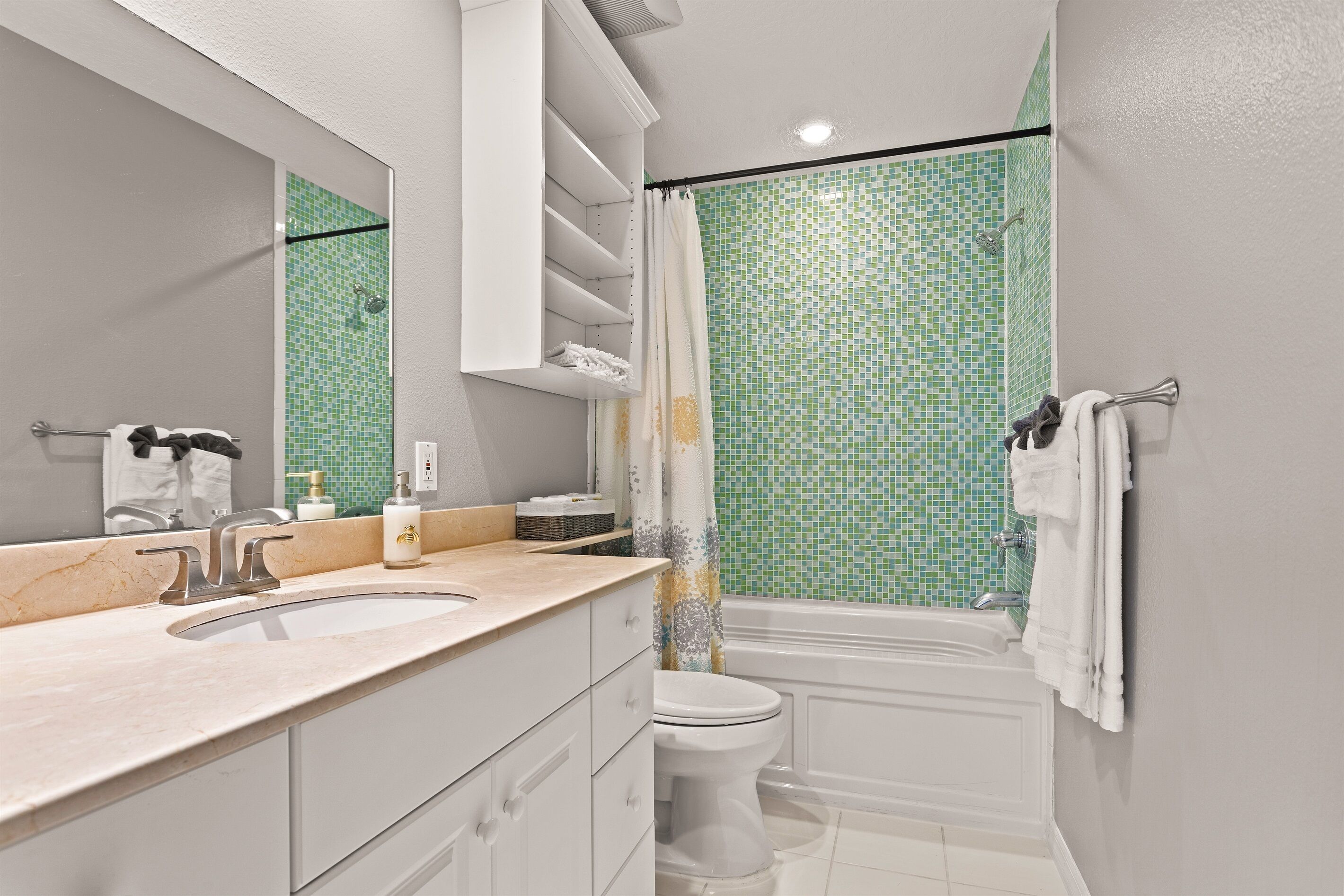 A modern bathroom with a large mirror, a sink, a toilet, and a shower with green mosaic tiles.