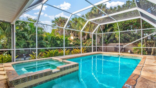 A swimming pool with a glass enclosure, surrounded by lush greenery and a wooden deck. - Next to IMG Golf! Heated Pool, Spa, Game Room! (Bradenton)