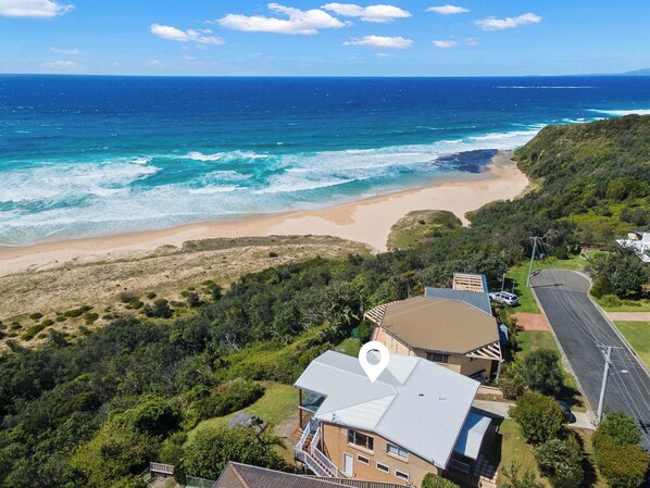 An aerial view of a coastal property with a beach, dense greenery, and a house with a visible roof and balcony.