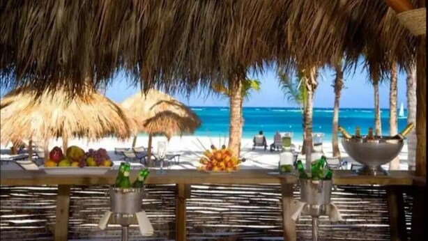 Eine Strandbar mit Reetdächern, Blick auf das Meer und einem Tisch mit Früchten und Getränken.