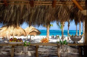Um bar à beira-mar com toldos de palha, vista para o oceano e uma mesa com frutas e bebidas.