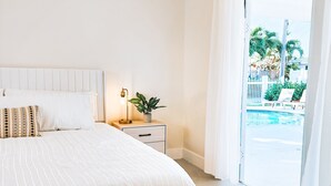 A bedroom with a large bed, a bedside table with a lamp, a potted plant, and a view of a swimming pool through a glass door.