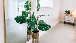 A modern interior with a large mirror, potted plants, a white dresser, and a window with blinds.