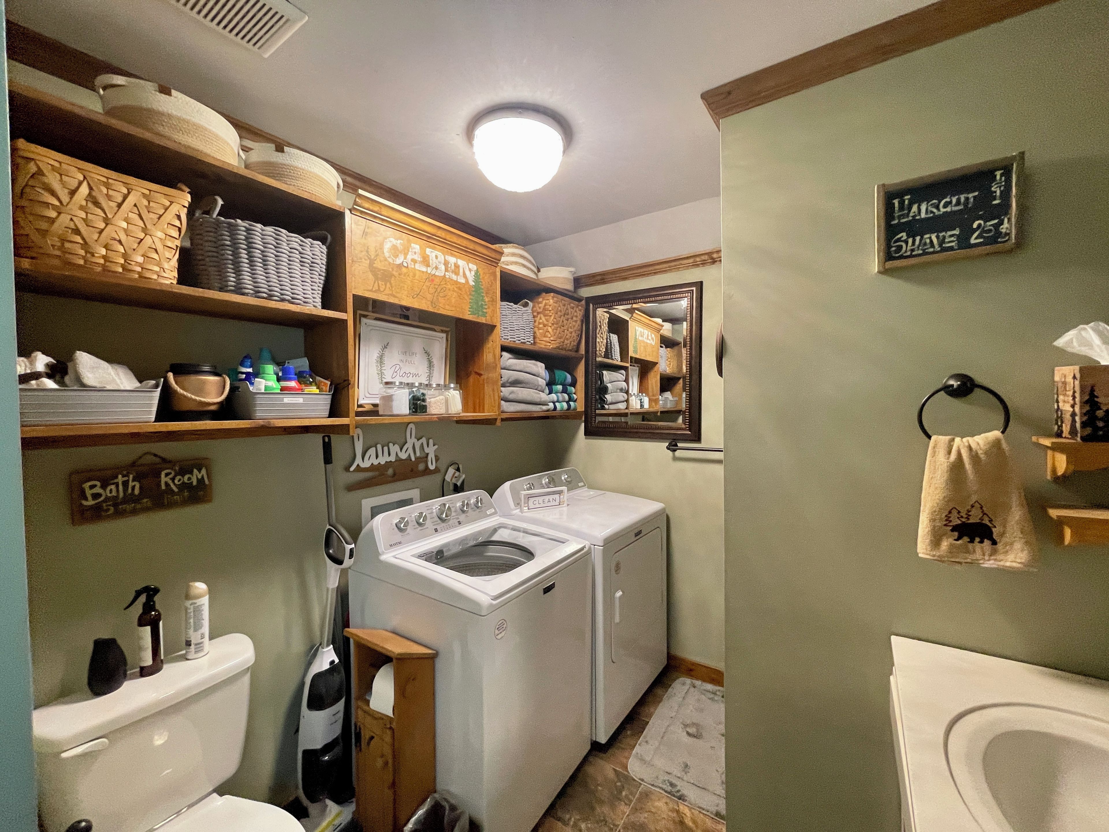 Baño pequeño y funcional con inodoro, lavadora y espejo. Se observan estantes con toallas y un cartel que indica 'baño'.
